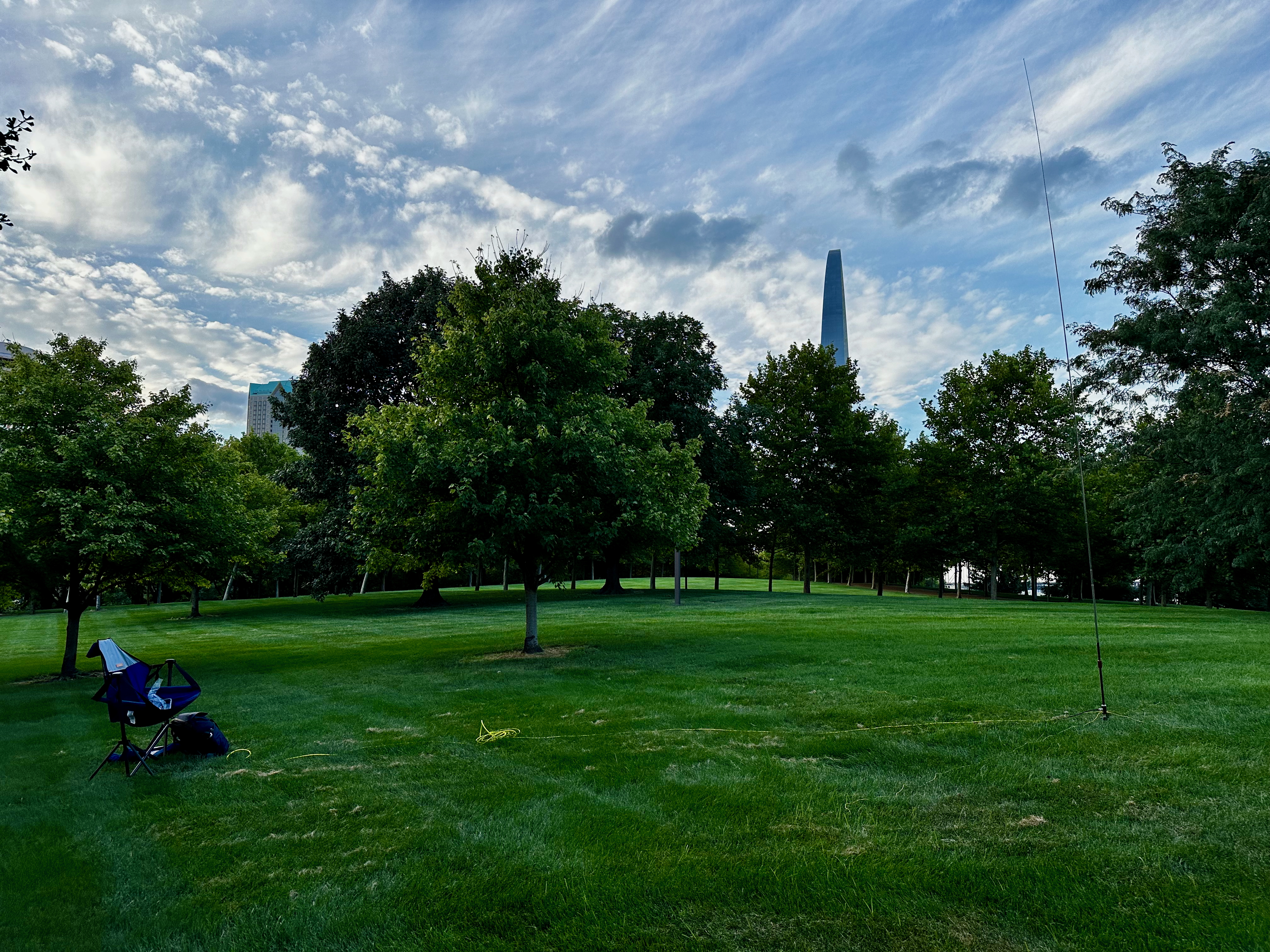 A small patch of grass amongst trees, the top of the Arch visible in the background, a camp chair with a radio and vertical antenna in the foreground.