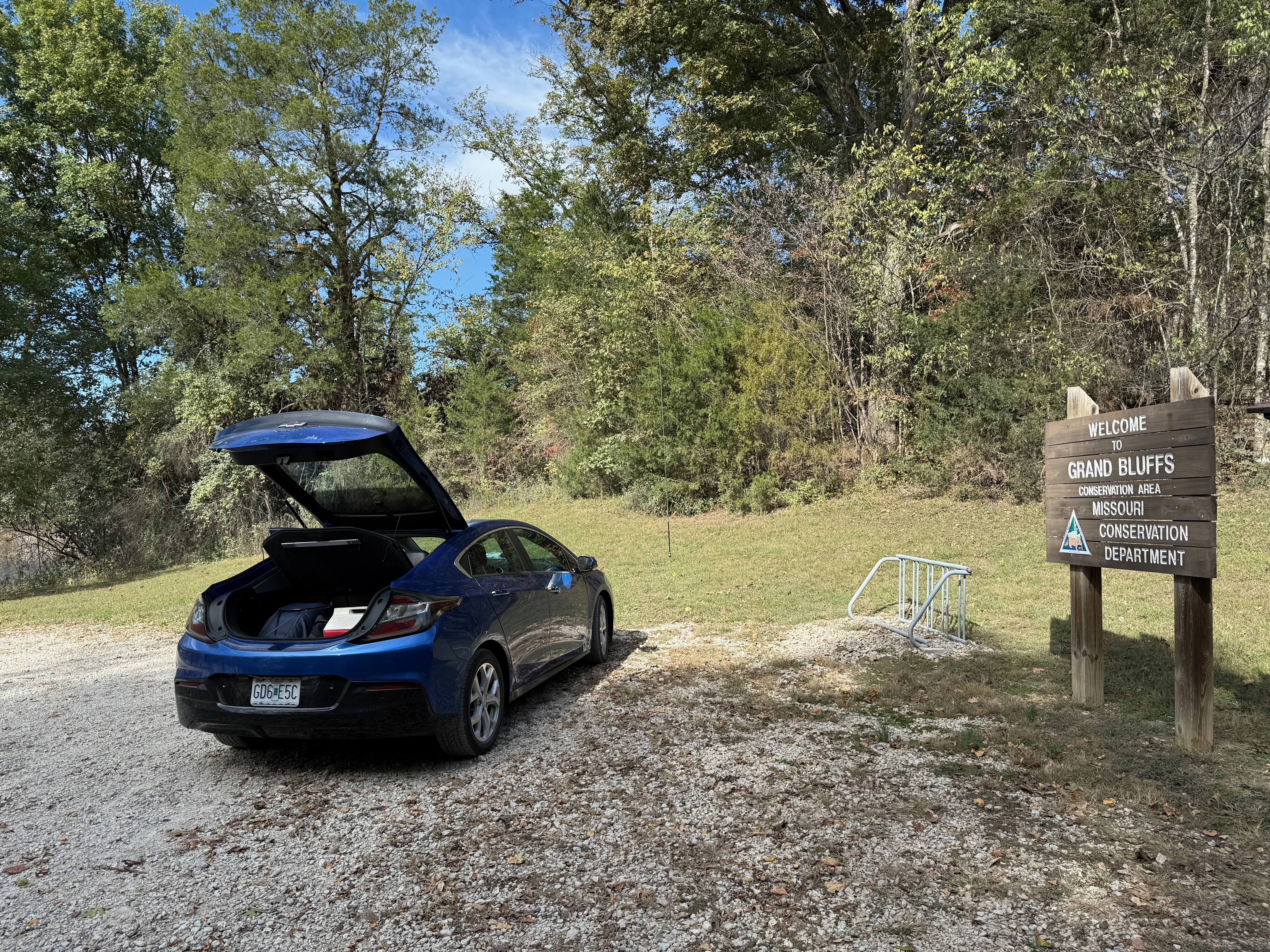 A gravel parking lot surrounded by green trees with a blue sedan with the trunk/hatch open on the left and a vertical antenna in the background in front of the car. On the right is a wooden sign that reads 'WELCOME TO GRAND BLUFFS CONSERVATION AREA MISSOURI CONSERVATION DEPARTMENT' and a small bike rack next to it.