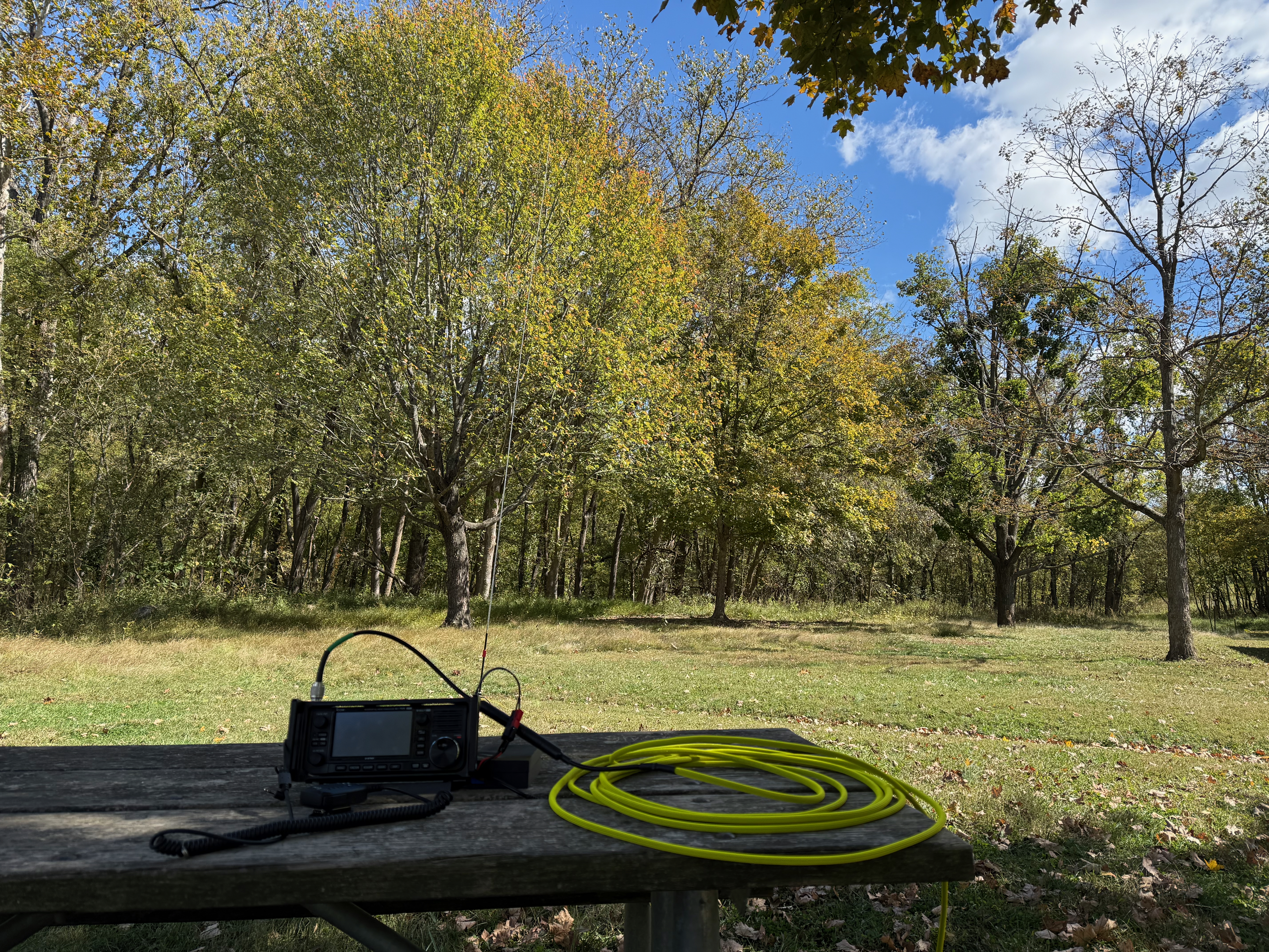 The right half of a shaded park bench with an Icom IC-705, a Bioenno battery and a few loops of yellow coax on it. The background is covered with green trees and has a small patch of mowed grass with a vertical antenna between the trees and picnic table.