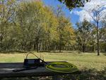 The right half of a shaded park bench with an Icom IC-705, a Bioenno battery and a few loops of yellow coax on it. The background is covered with green trees and has a small patch of mowed grass with a vertical antenna between the trees and picnic table.