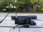 A park bench with an Icom IC-705, microphone, Bioenno battery and carrying case for the Chelegance MC-750 antenna. A short length of black coax runs off to the left. The Katy Trail, some trees and an old concrete building is in the background.