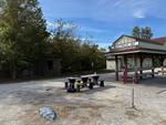 A wider shot of the park bench on some gravel. On the right in the foreground in the vertical antenna. In the background is the McKittrick, MO trailhead with a small shelter intended to vaguely resemble an old train station with some benches and informational display.