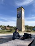 A view of the park bench from the other side with me sitting facing the camera. An old conrete gain silo with an aerial view of the Mississippi River is painted on two sides with 'Magnificent Missouri' written immediately below.