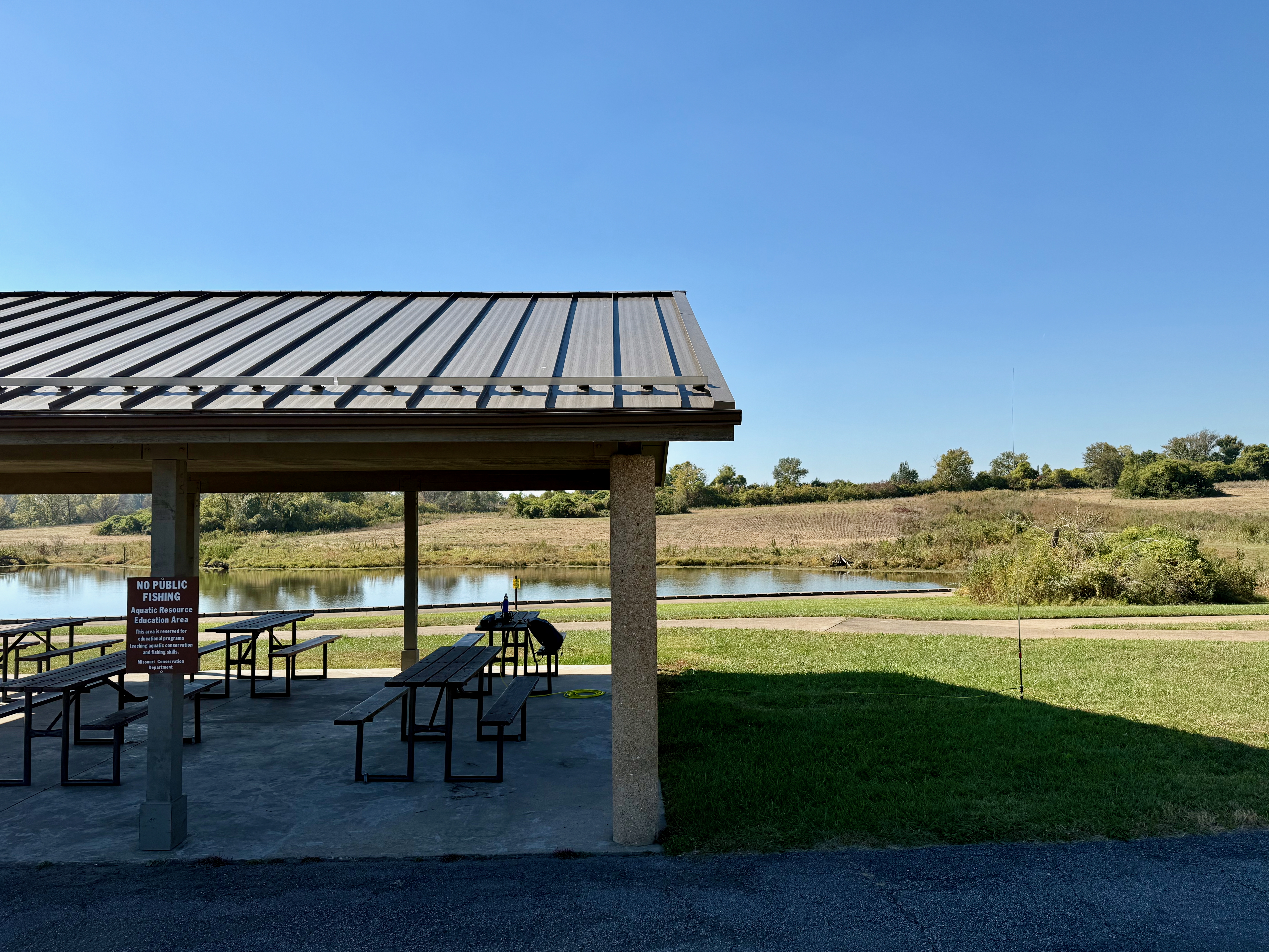 One half of a large park shelter with park benches. The furthest park bench contains some radio equipment, a backpack and yellow coax running to a vertical antenna to the right. A large pond in the background with a grassy plain and some trees behind that under a clear blue sky.