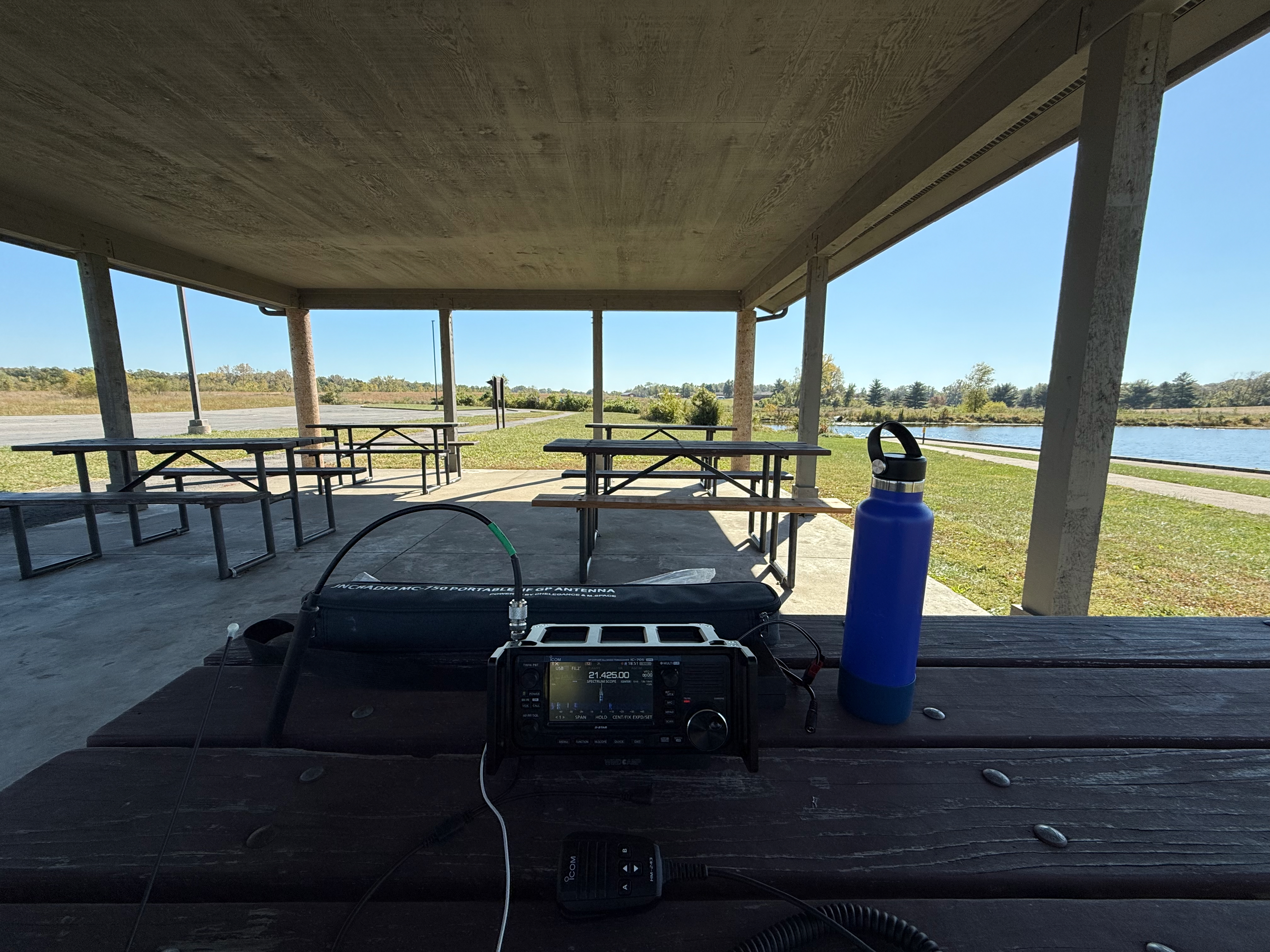 A closer view of the picnic table with the radio equipment on it under the park shelter. An Icom IC-705, hand mic, a Chelegenace MC-750 carrying bag and a water bottle are on the table.