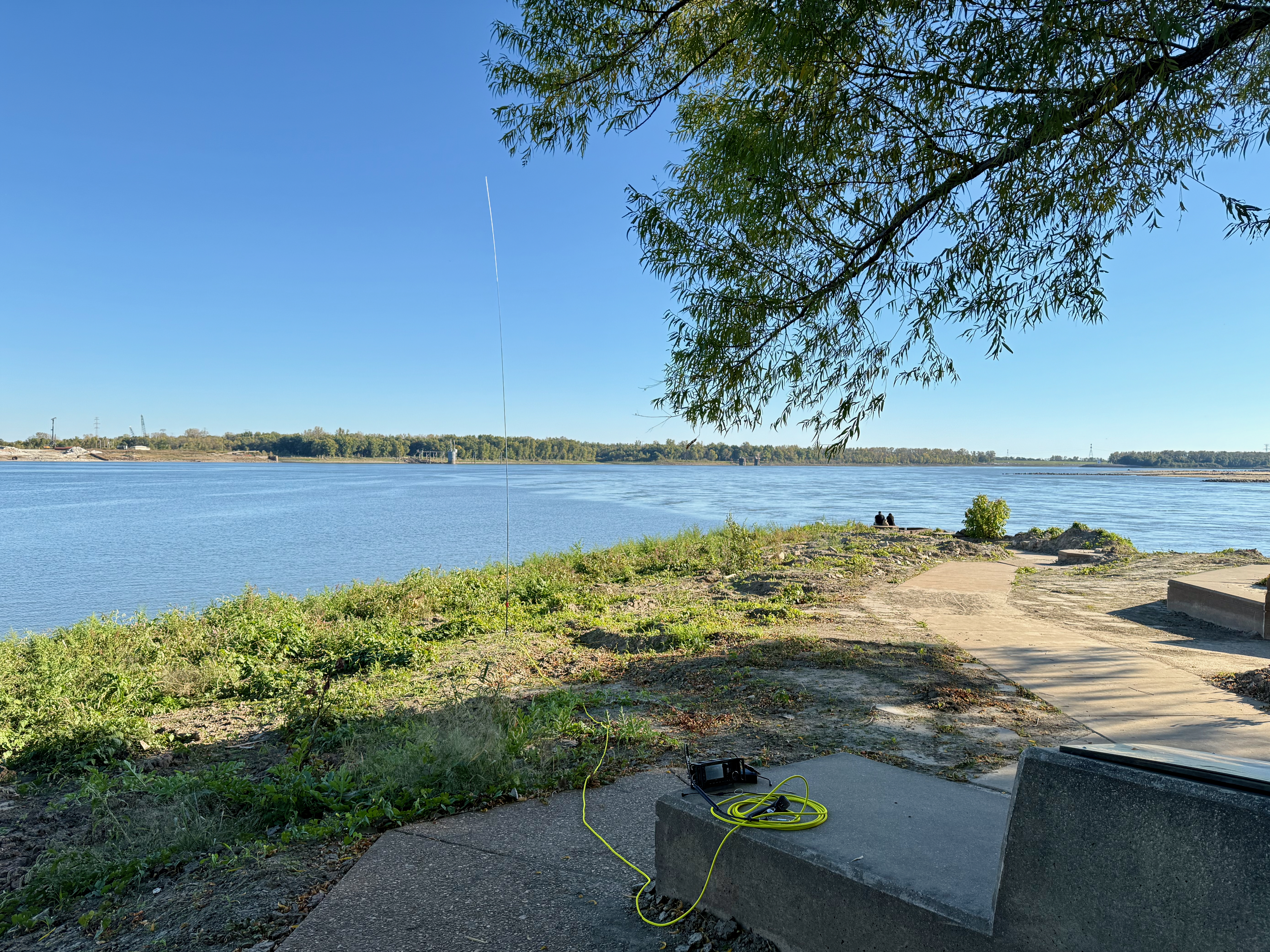 A wide view of the confluence of the Mississippi and Missouri river from about 50 feet back from the shores. A large tree to the right side of the image provides shade for an Icom IC-705 radio on a large concrete slab. Yellow coax snakes off to a vertical antenna closer to the shore. A couple sits at the edge of the shore near the confluence.