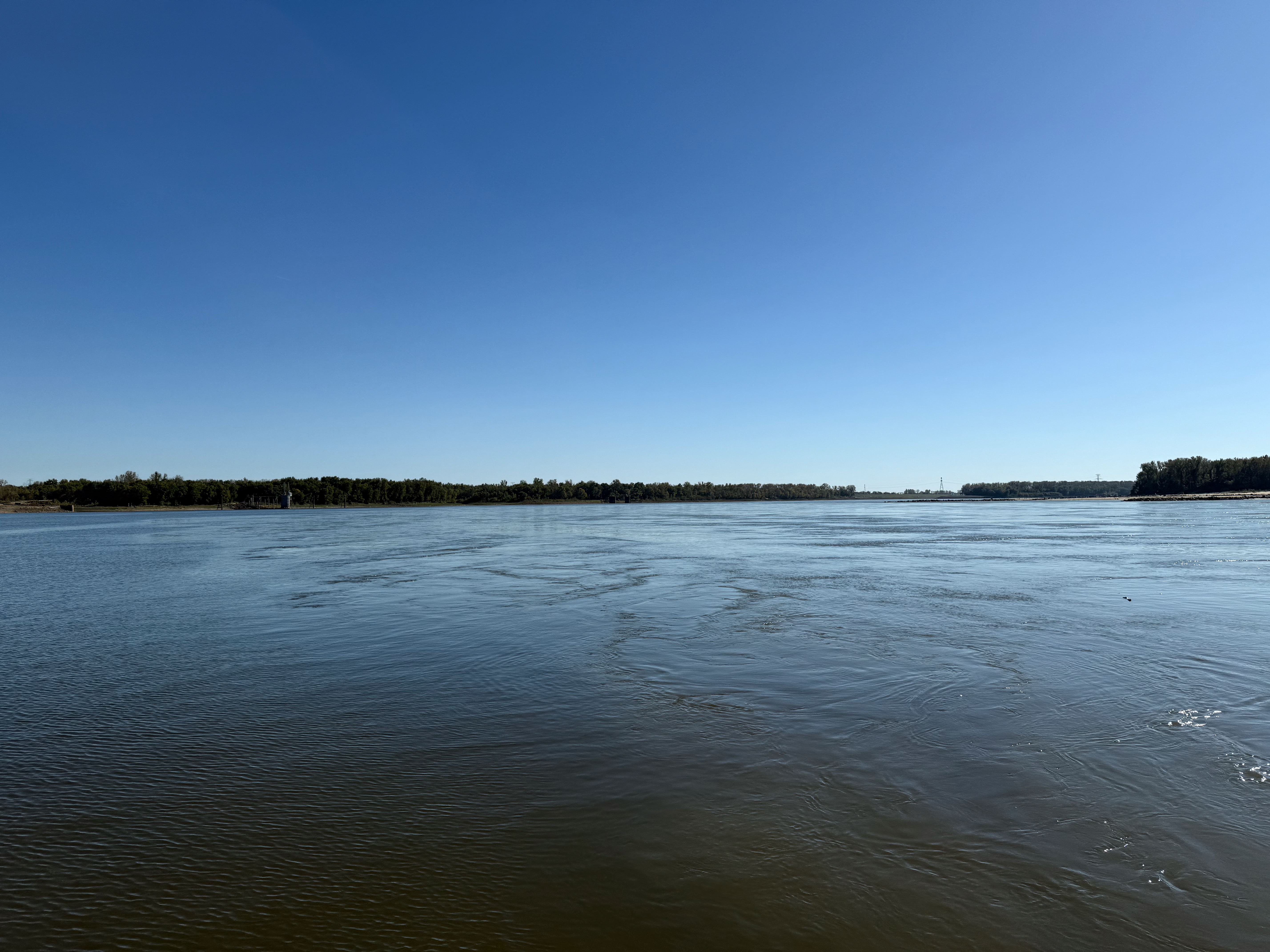 A closer view of the river confluence under a clear blue sky with trees in the distance on the Illinois side of the river. The calmer Mississippi river is on the left and the muddier Missouri river on the right. The area where the two rivers meet is more turbulent and rough.