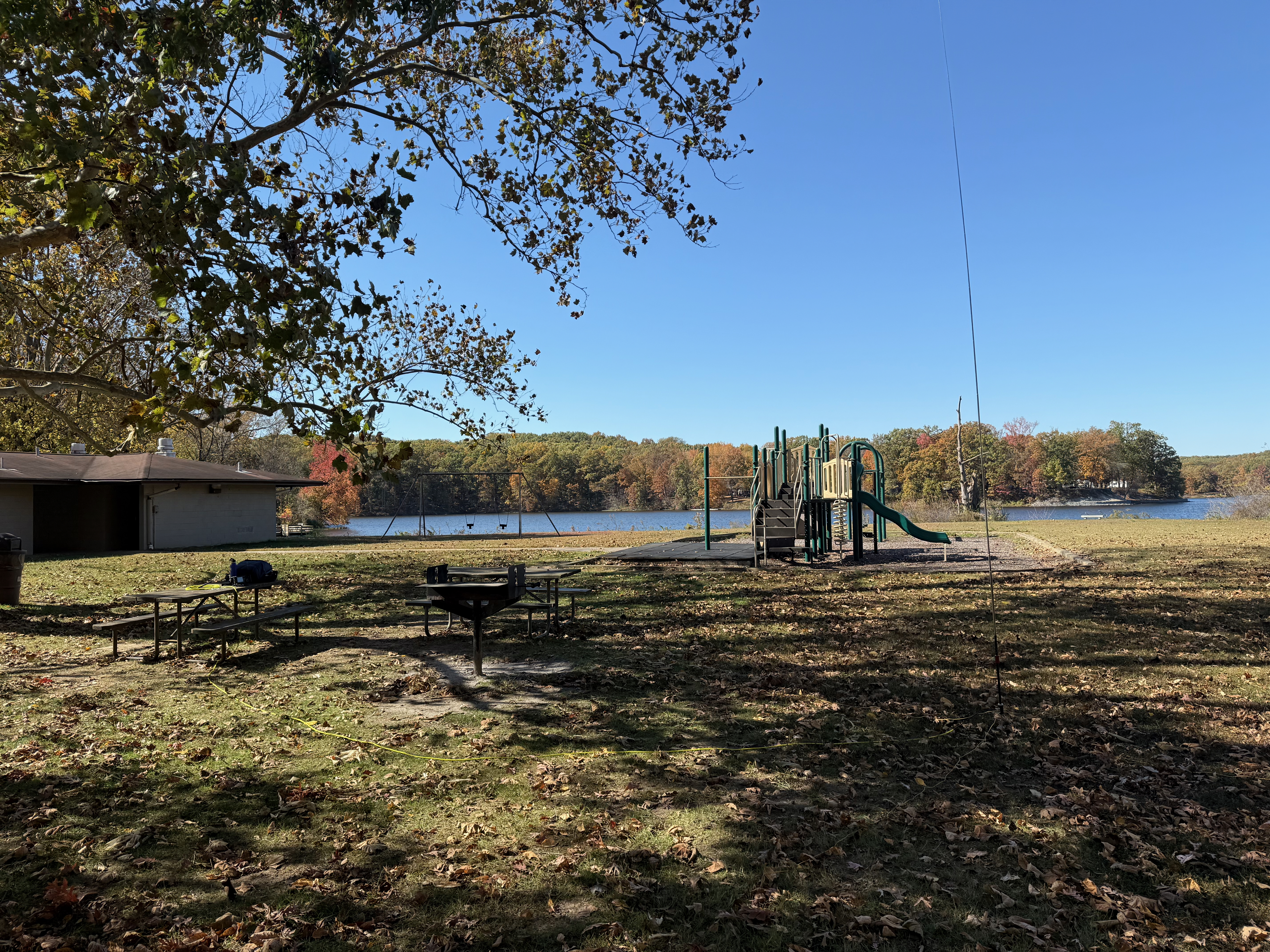 The area near the lake I activated from. A park bench and grill under a tree on the left with the vertical antenna on the right. In the background is a concrete building housing the bathrooms on the left and a small playground in the middle.