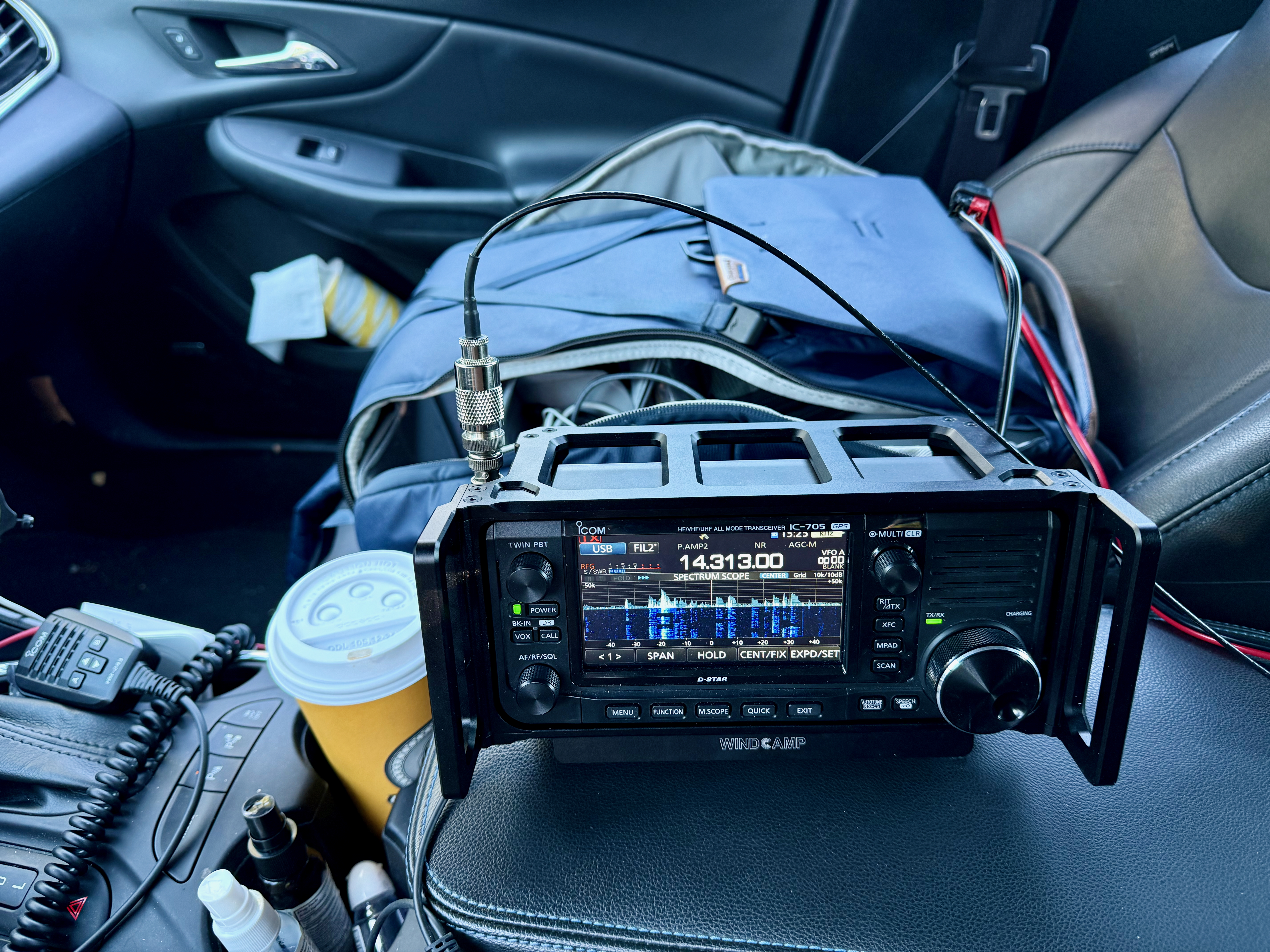 A view from inside a car showing an Icom IC-705 radio on the center arm rest with a backpack in the seat behind. The hand mic is to the far left and a large paper coffee cup in a cup holder.