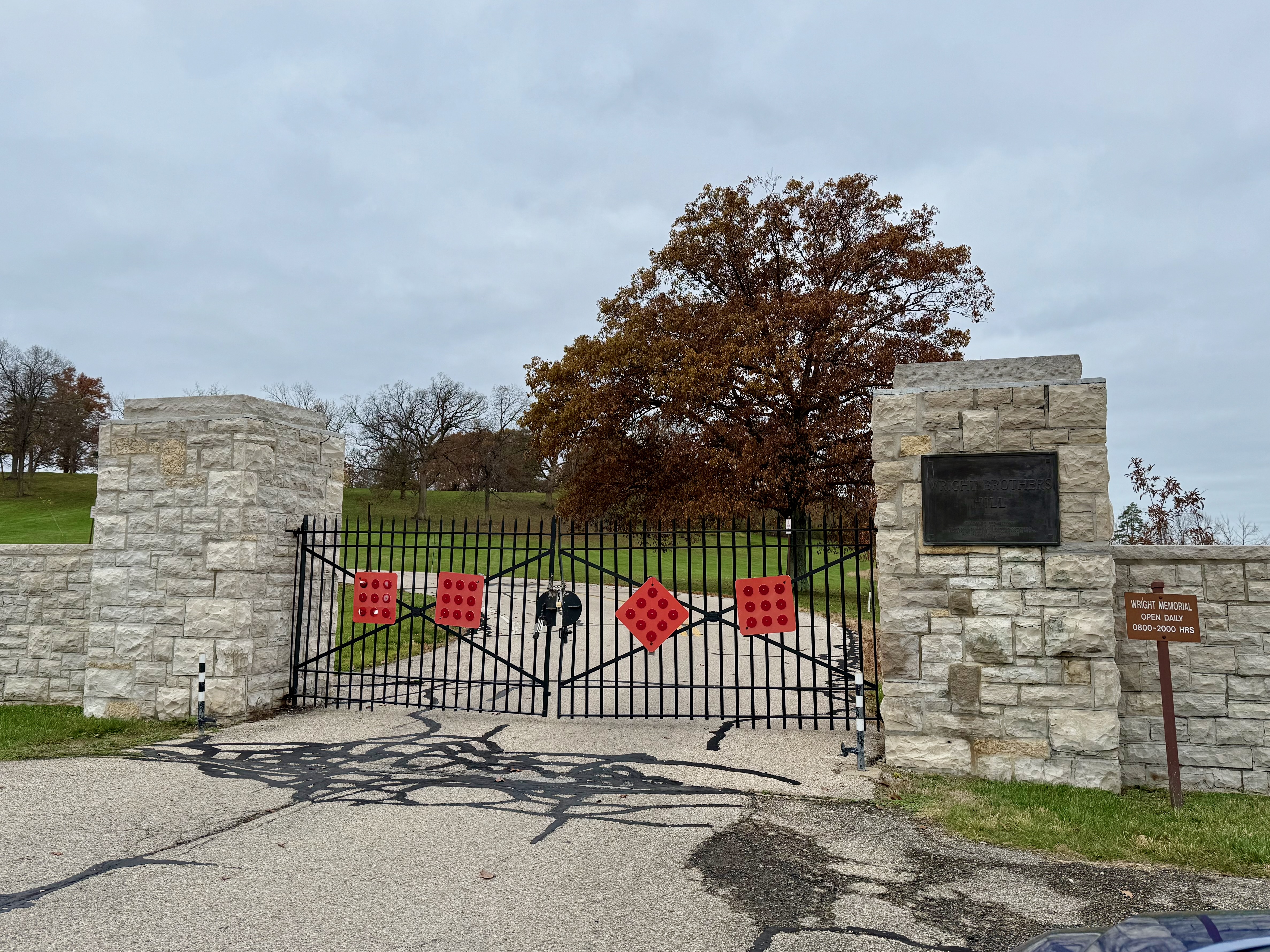 The locked wrought iron gate to the park between two sandstone pillars. A sign on the right reads 'Wright Memorial Open Daily 0800-20000 HRS'