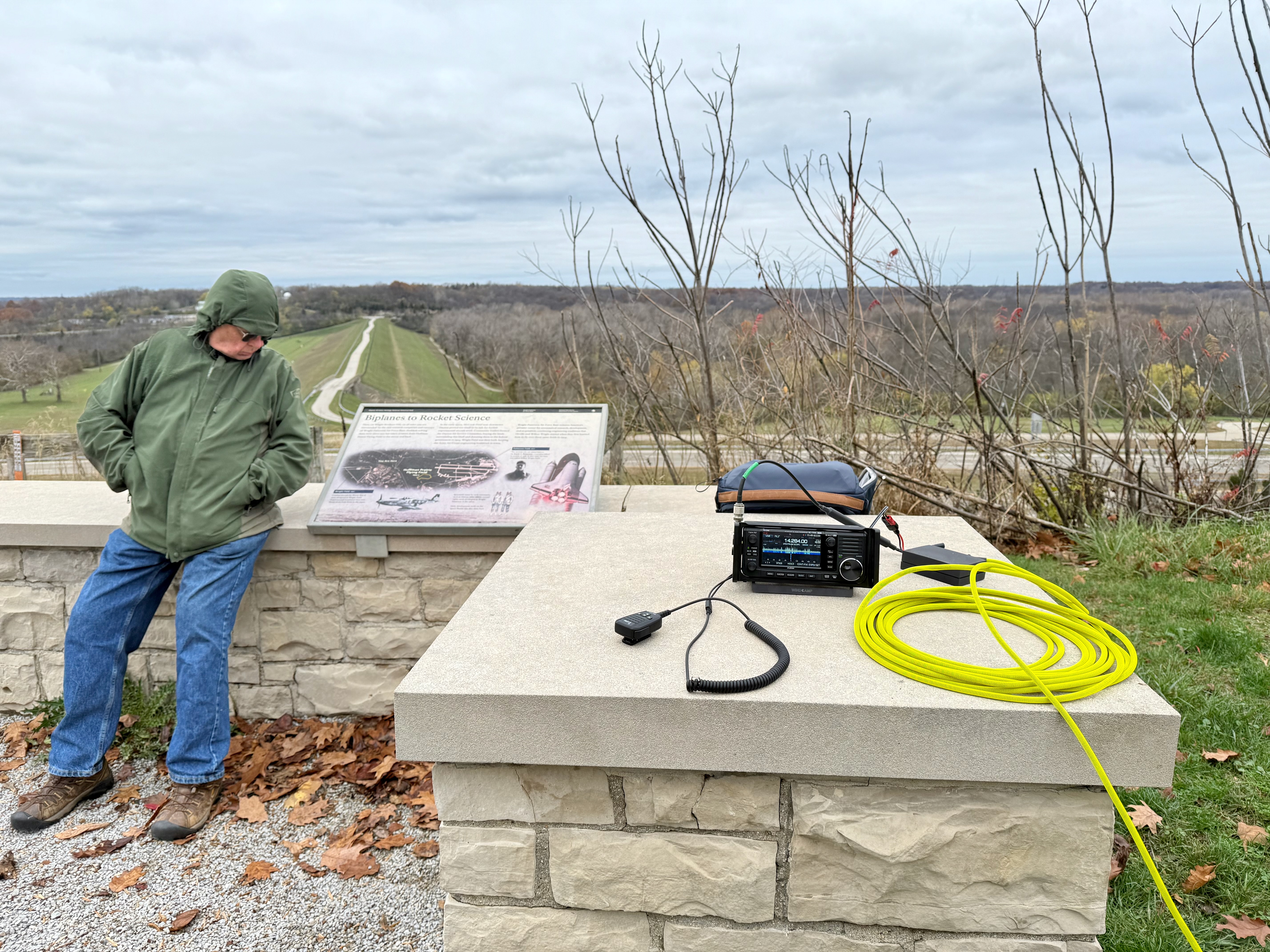 A view of the setup. A radio, microphone, battery and coax is sitting on a sandstone wall on the left in the foreground. In the distance is a slightly elevated view of the horizon to the north. My Dad is sitting on the wall to the left next to an informational sign.