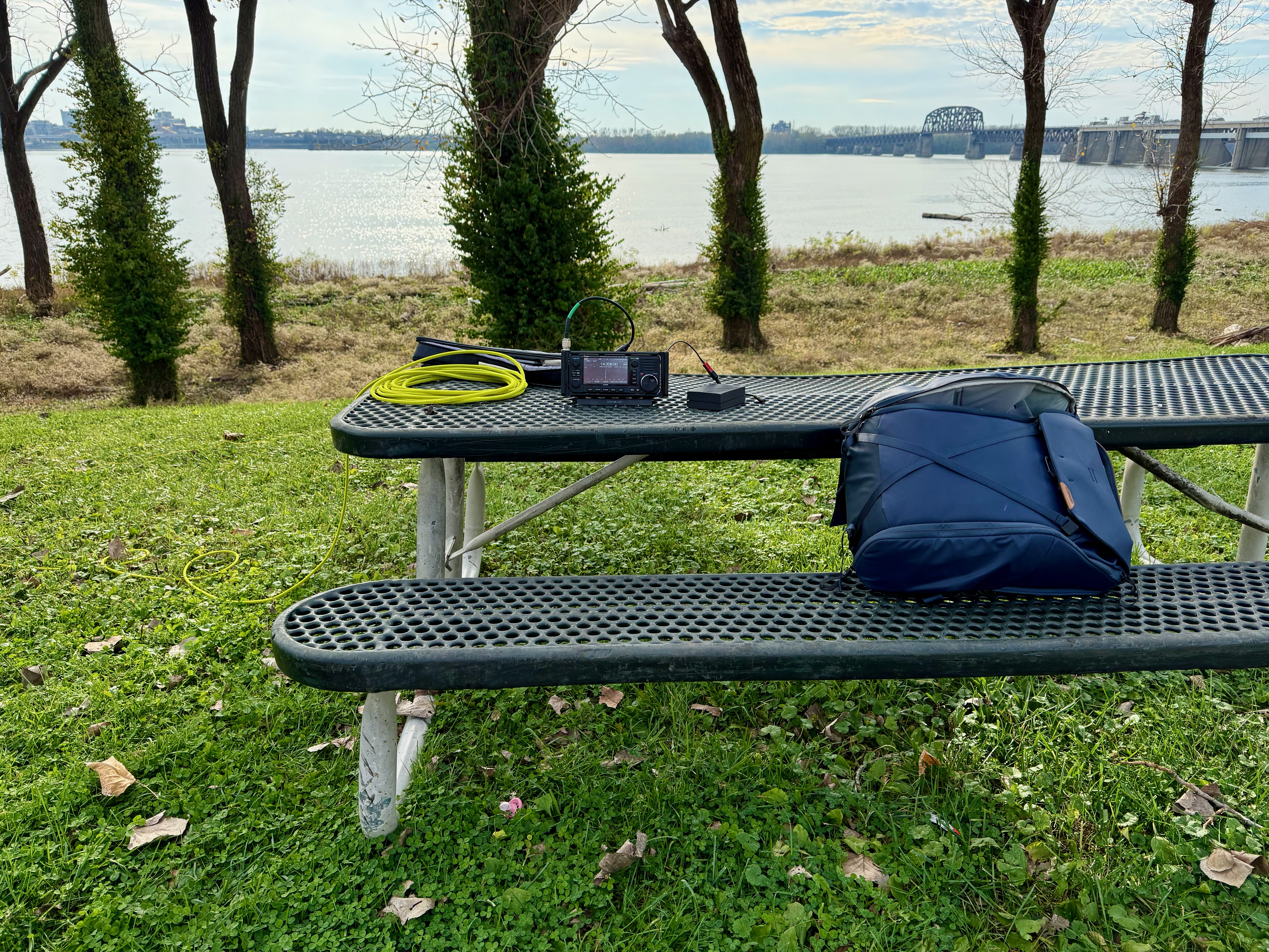 My radio equipment on a picnic table at the park. In the background is the Ohio river with some bare trees between the picnic table and river.