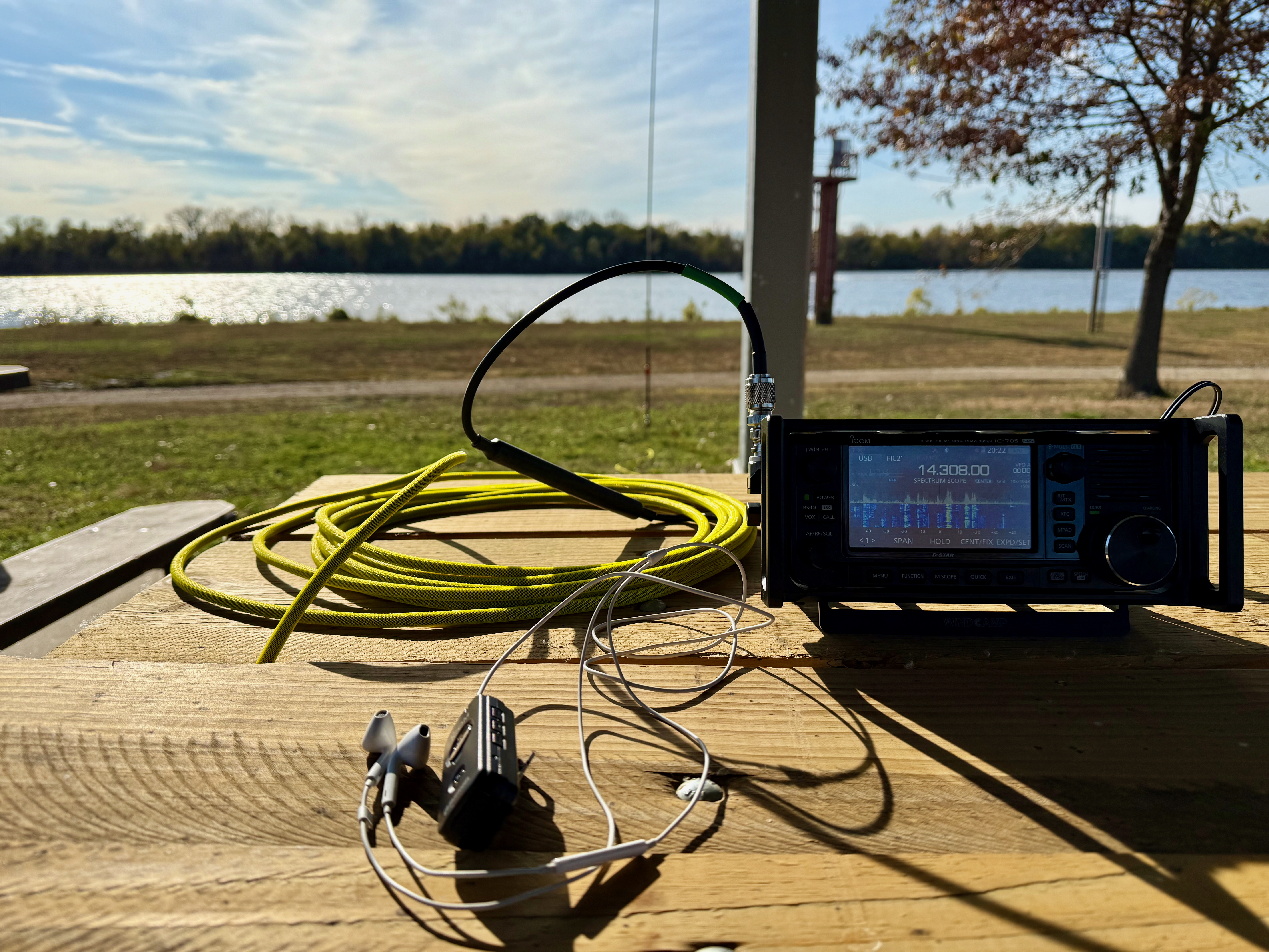 The setup on the picnic, the sun casting long shadows on everything. An Icom IC-705, hand mic, yellow coax and carrying case for the Chelegance MC-750 in the foreground. The Mississippi River is in the background.