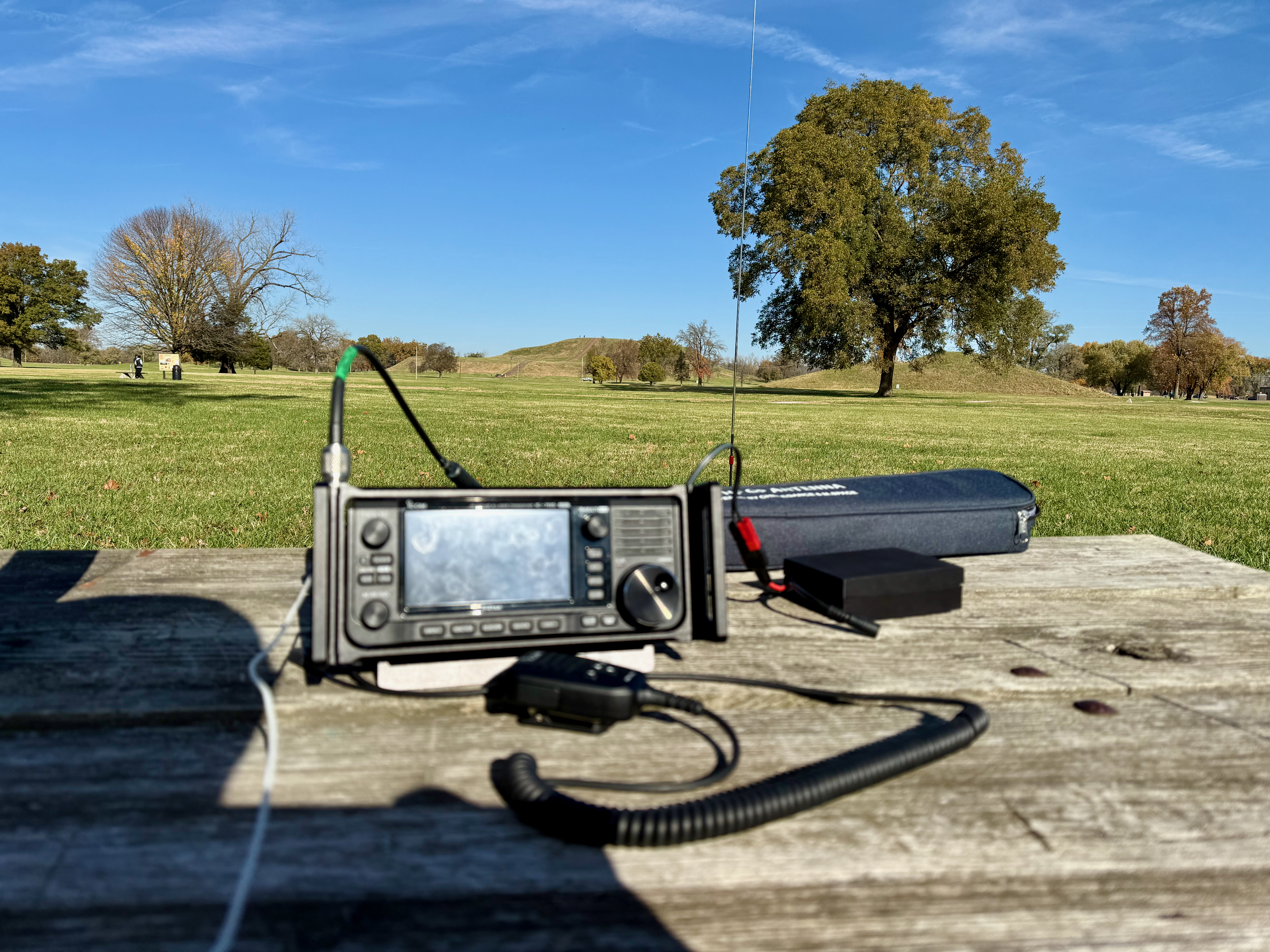 A partially-shaded picnic table under clear blue skies. An Icom IC-705, microphone and Bionneo battery are on the table, a vertical antenna just behind that. In the distance Monk's Mount is visible, rising gently up.