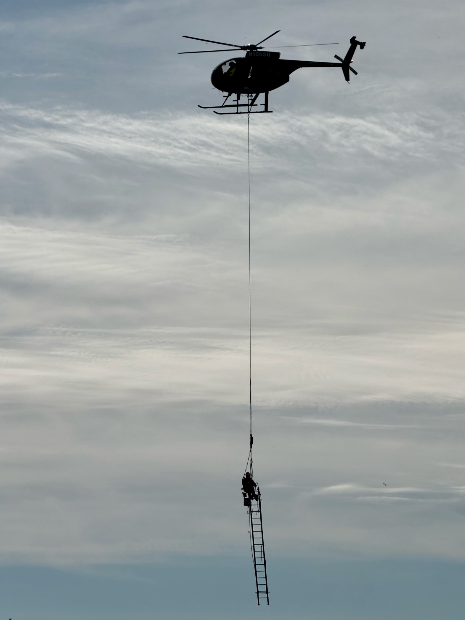 The helicopter in the sky with a line down a ladder with someone at the top of the ladder.