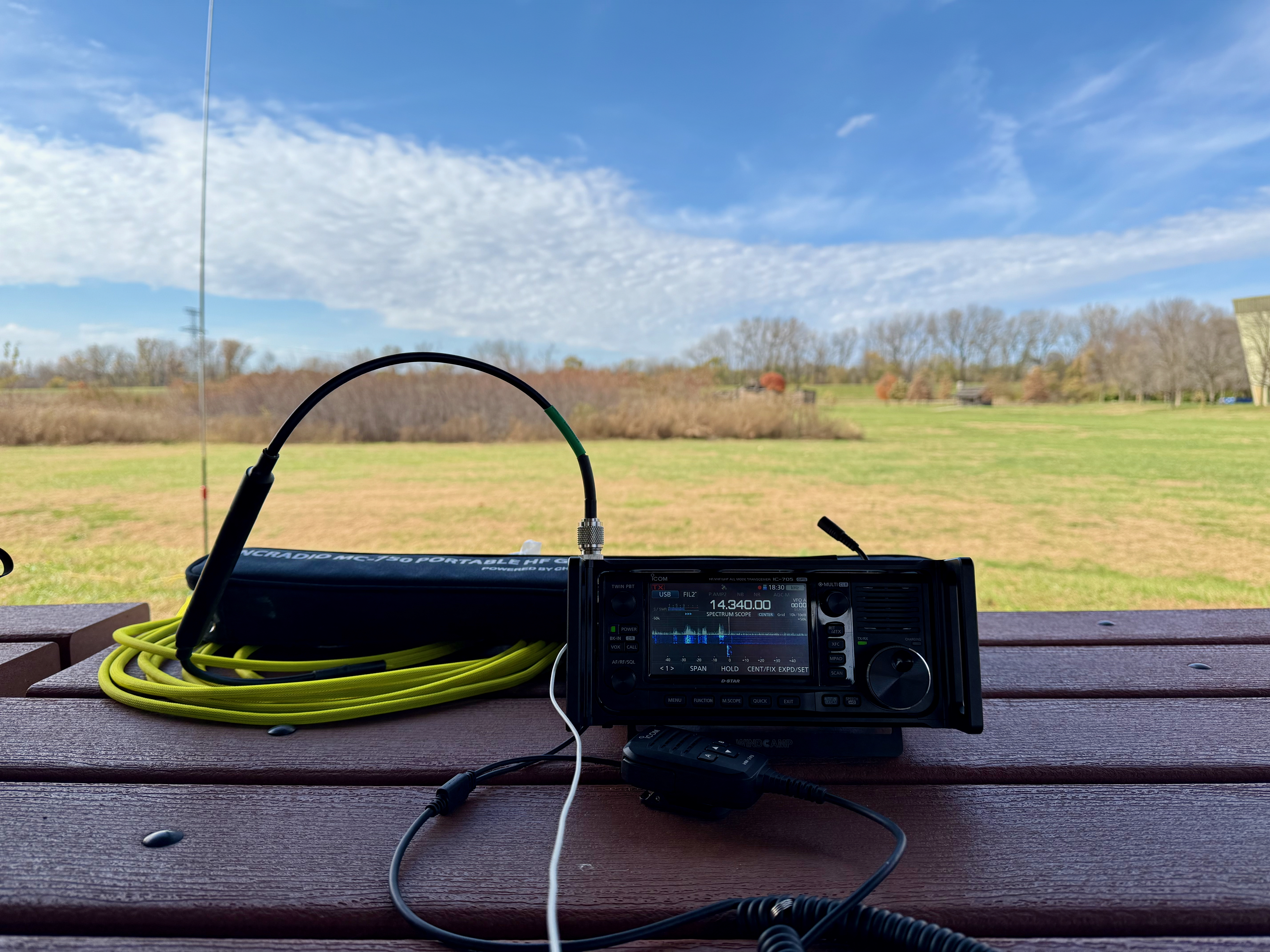 The setup on the picnic table under the shade of the shelter. An Icom IC-705, hand mic, yellow coax and carrying case for the Chelegance MC-750 in the foreground. A grassy field under a party cloudy sky in the background.